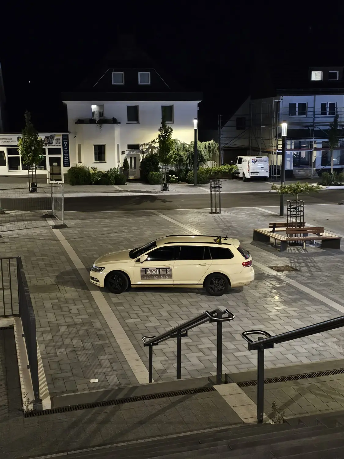 BonChance & TaxiTopTeam GmbH - Transportdienste - Ein weißes Taxi parkt nachts auf einem Stadtplatz oder einer Straße; im Hintergrund sind Gebäude und Treppen zu sehen.