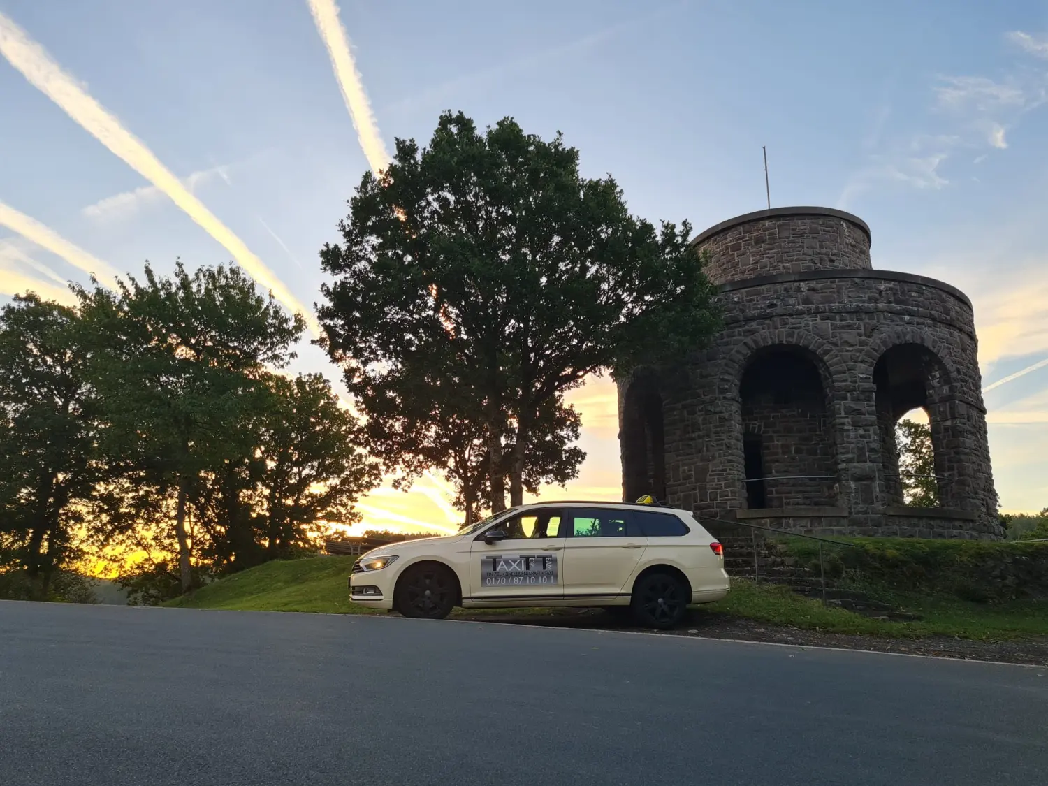 BonChance & TaxiTopTeam GmbH - Transportdienste - Bei Sonnenuntergang parkt ein weißes Taxi neben einem Steinturm und großen Bäumen.